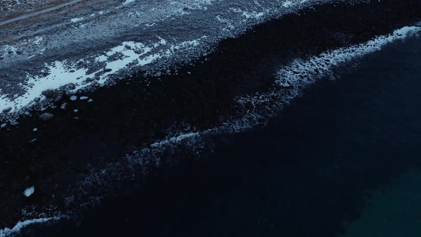 A mesmerizing top-down aerial view of dark arctic waves gently lapping against a snowy, rocky shoreline