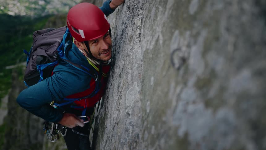 A male rock climber in a helmet smiles at the camera before clipping a quickdraw into a bolt on a high mountain cliff face