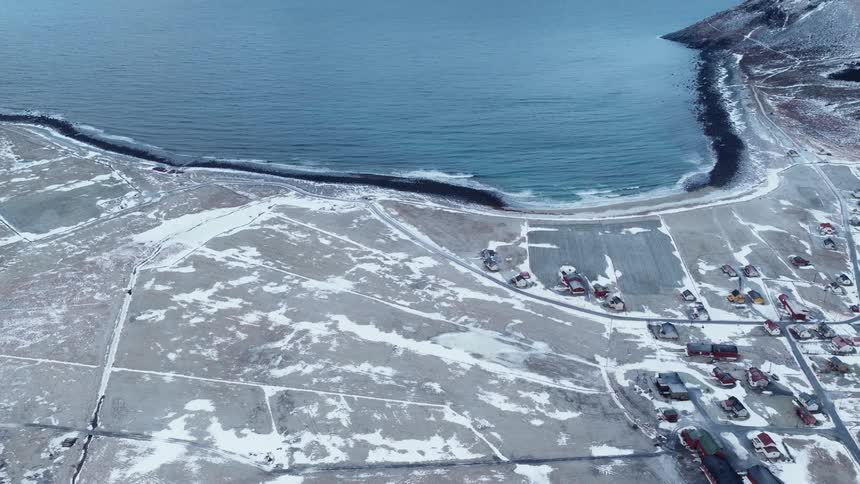 High angle aerial shot tilts up to reveal a stark and beautiful snowy coastline with a small village and vast ocean