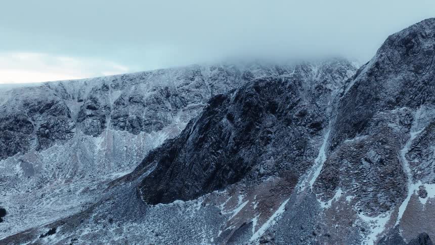 A slow and majestic aerial reveal of a vast, snow-dusted mountain range with its peaks shrouded in clouds