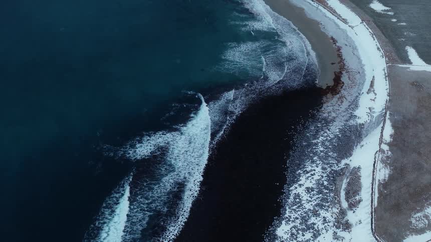 An aerial top-down shot slowly descends over a dramatic, snow-covered coastline as dark ocean waves crash against the shore