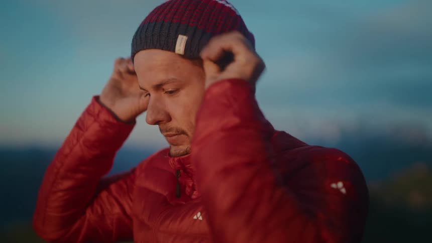 A man zips up his red puffer jacket and puts on a beanie, preparing for the cold in the mountains at dusk