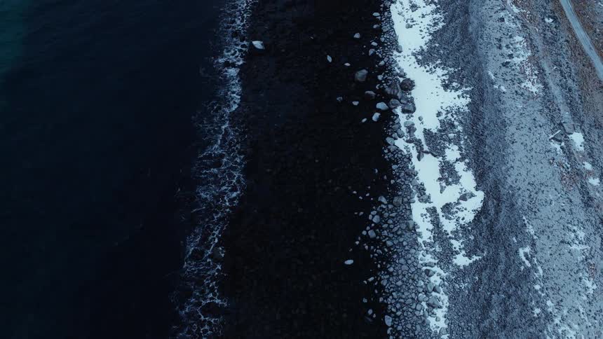 A slow aerial view follows the dark, cold ocean waves washing over a rocky shoreline partially covered in snow