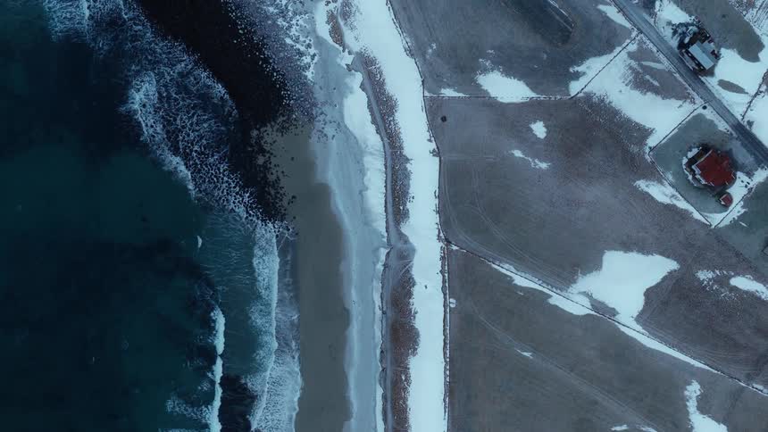 Top-down aerial shot slowly rises over a stark winter coastline where the dark ocean meets a snow-covered beach and rural landscape