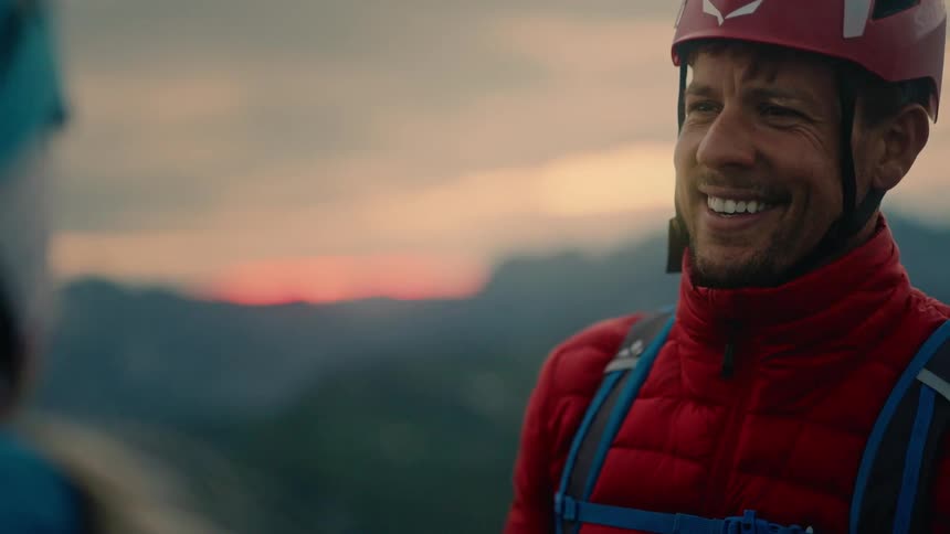 A smiling male climber gives a celebratory handshake to his partner after reaching a mountain summit at sunset