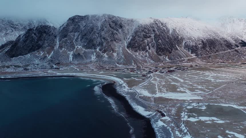 An epic aerial view captures a remote coastal village nestled between the dark sea and a majestic snow-capped mountain range