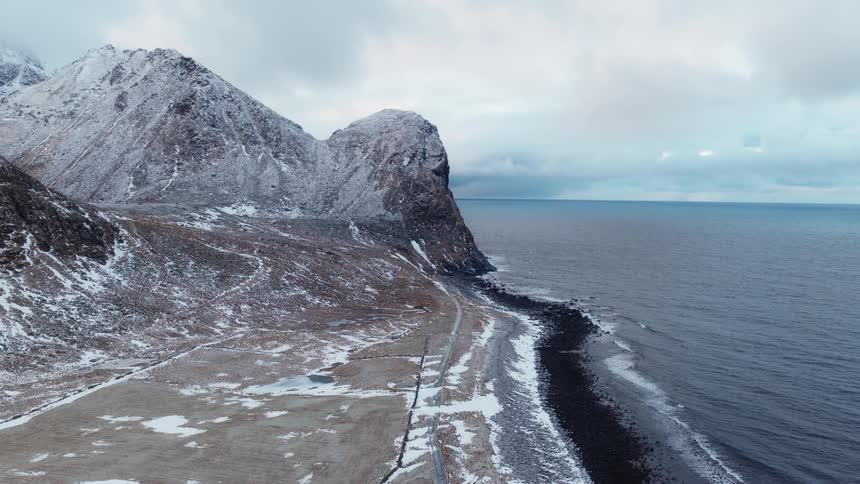 A dramatic aerial view of a snow-covered mountain range towering over a rocky coastline on a cold, overcast day