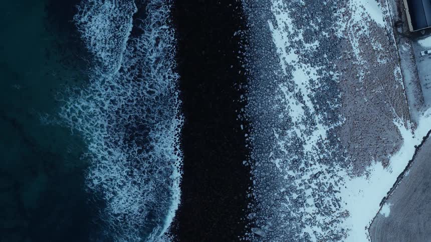 A dramatic top-down aerial view reveals dark ocean waves crashing onto a frozen, snow-dusted shoreline