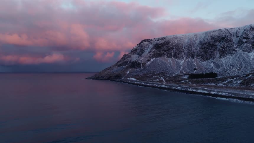 A dramatic aerial view shows a snowy mountain coastline meeting the calm ocean under a vibrant pink and purple winter sunset