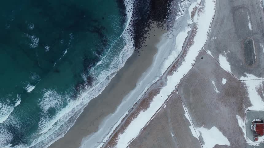 A stunning top-down aerial view captures the contrast of turquoise ocean waves gently lapping against a snow-dusted shoreline