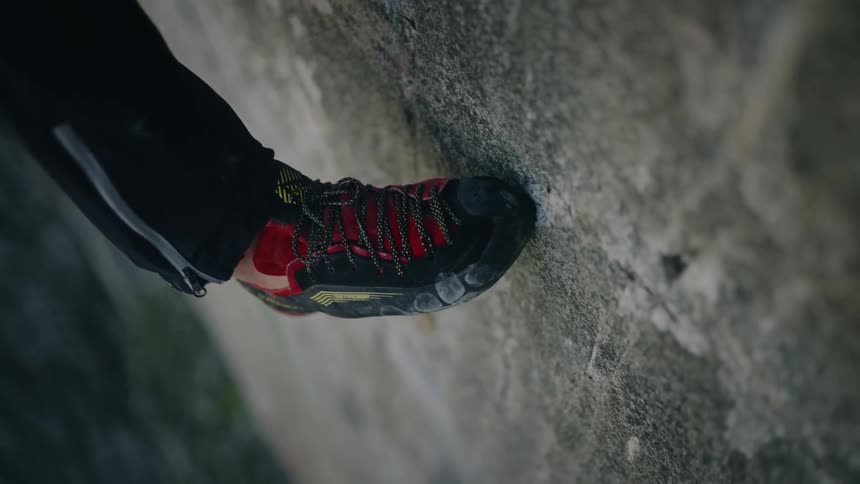 A detailed vertical shot shows a rock climber's specialized shoe carefully finding its grip on a rock in slow motion