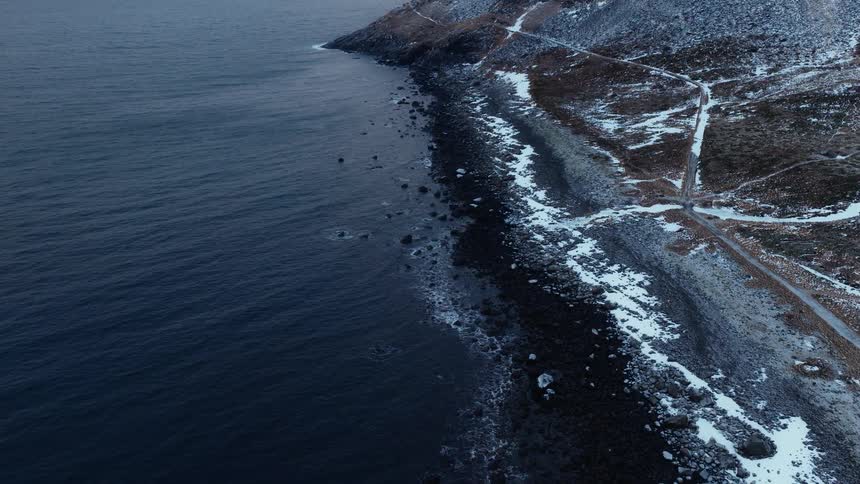 An aerial view captures the moody and desolate beauty of a snowy, rocky coastline as dark ocean waves gently wash ashore