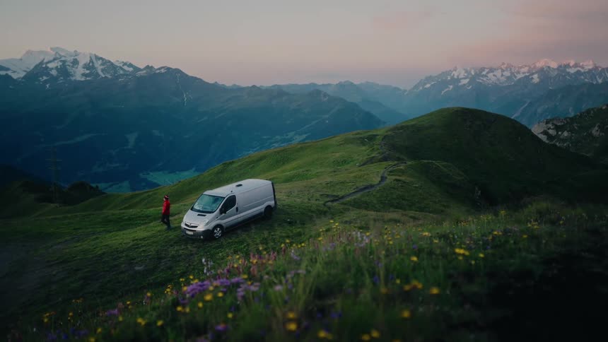 An adventurous person gets out of their camper van to enjoy the stunning sunrise view over the snow-capped mountains from a grassy, wildflower-covered hill