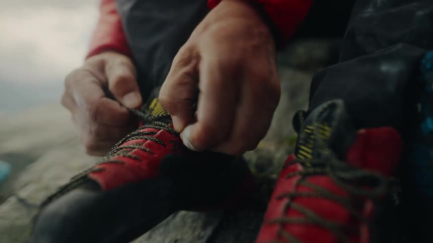 A close-up of a rock climber's hands tying the laces on his red climbing shoes before an ascent