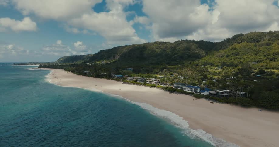 A static aerial view of a picturesque tropical coastline with turquoise water, a sandy beach, and lush green mountains under a partly cloudy sky