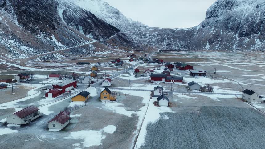 A scenic aerial shot flies from a snow-dusted beach toward a remote coastal village nestled in a valley between towering winter mountains