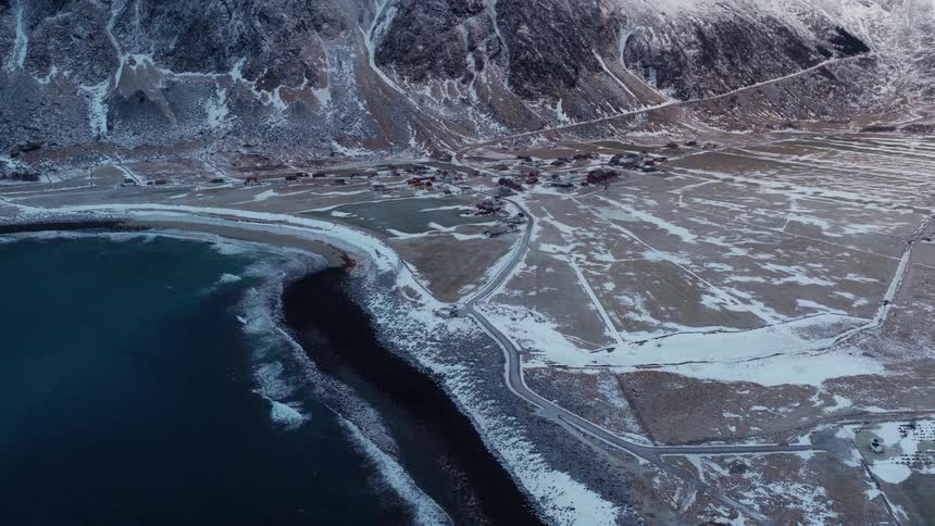 A stunning aerial reveal of a remote coastal village at the base of majestic snow-covered mountains in winter