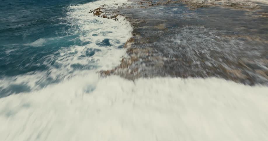 A dynamic aerial shot flies low over powerful ocean waves as they crash onto a rocky tropical shoreline under a partly cloudy sky