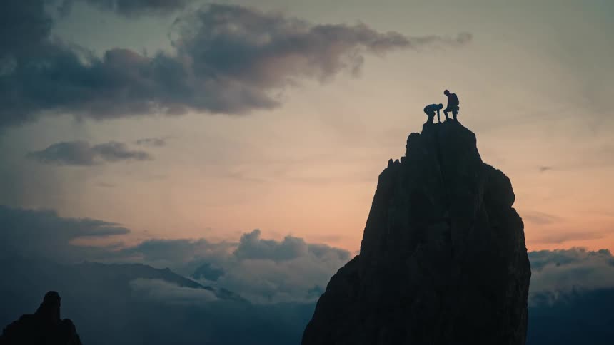 Two climbers reach the top of a mountain, where one helps the other up, and they celebrate their success with a high-five at sunset