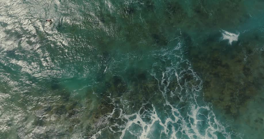 A stunning top-down aerial view captures a surfer paddling out into the turquoise sea before being swallowed by a powerful wave