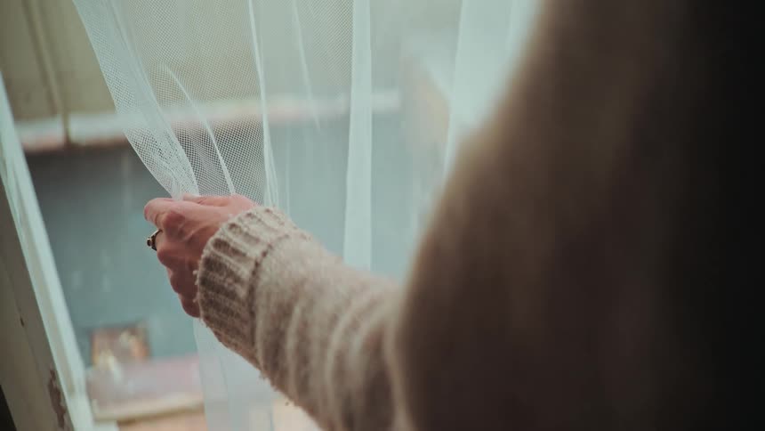 A person's hand in a cozy sweater slowly pulls back a sheer white curtain to look out the window