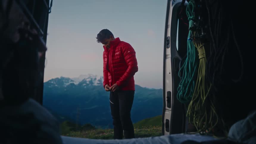 A male adventurer puts on a red puffer jacket by his camper van, preparing for a day in the stunning snow-capped mountains