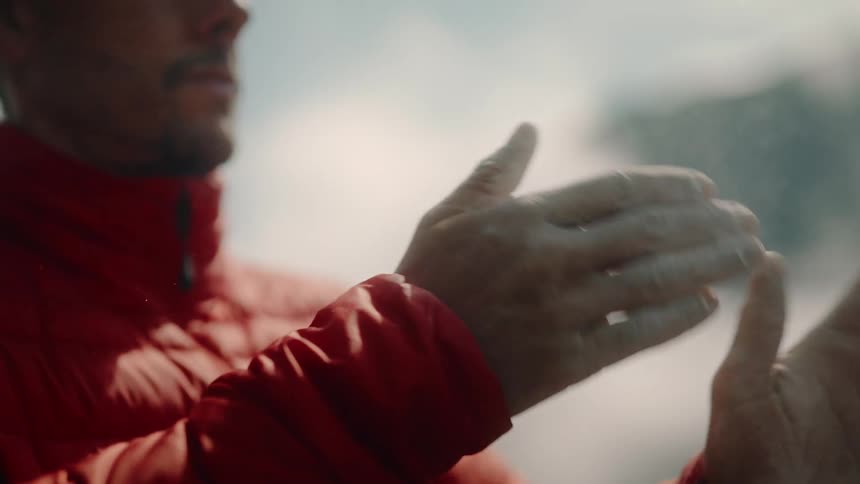 A climber in a red jacket applies chalk to his hands in slow motion, looking up with focus and determination before his ascent