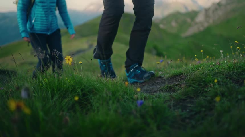A slow-motion close-up captures a couple's boots as they hike along a scenic green mountain path