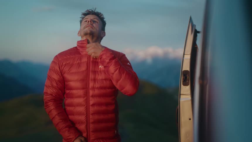 A smiling man puts on a warm red puffer jacket and beanie next to his camper van, enjoying the stunning mountain sunrise