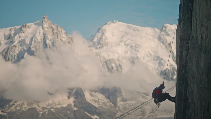 A lone climber rappels down a sheer rock face with a breathtaking view of snow-capped mountains in the background