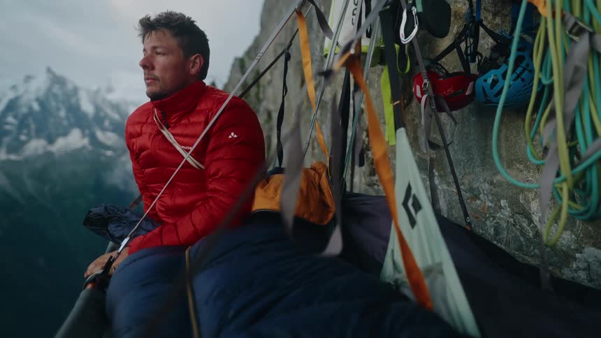 An alpinist wakes up in his portaledge on a steep mountain cliff and enjoys the stunning view