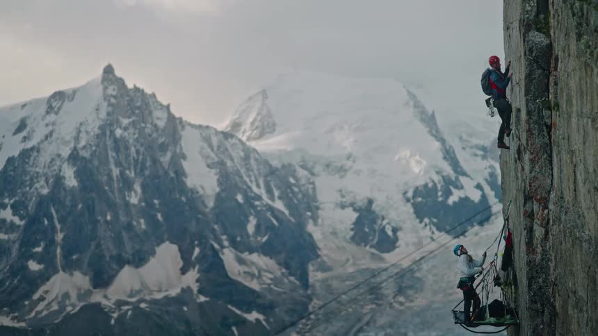 A rock climber carefully makes their way up a steep cliff face while another rests on a portaledge, with a majestic snow-covered mountain in the background