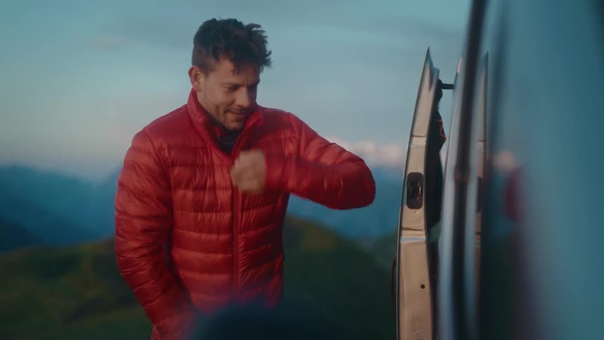 A man prepares for an early morning adventure by putting on a warm red puffer jacket and beanie next to his van in the mountains