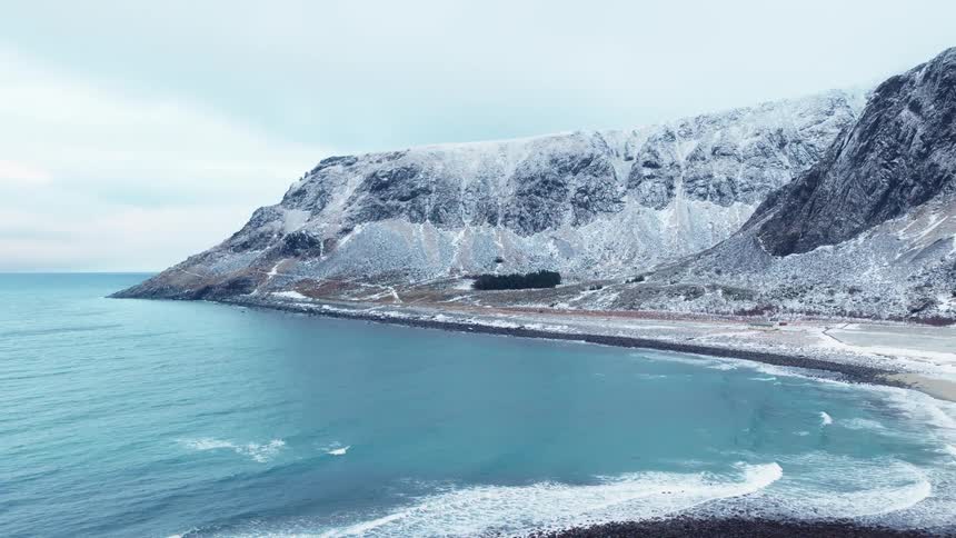Ascending aerial view of a stunning winter coastline with turquoise waves washing ashore on a pebble beach at the base of snow-covered mountains