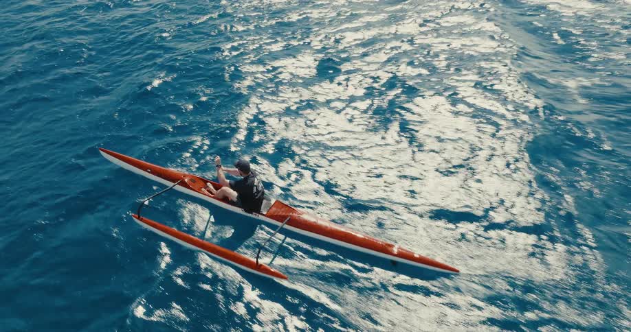 An aerial shot follows two men as they paddle their outrigger canoes across the vast, sun-kissed blue ocean