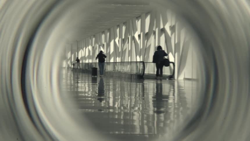 A stylized, desaturated view through a circular frame shows travelers with luggage walking down a modern airport hallway