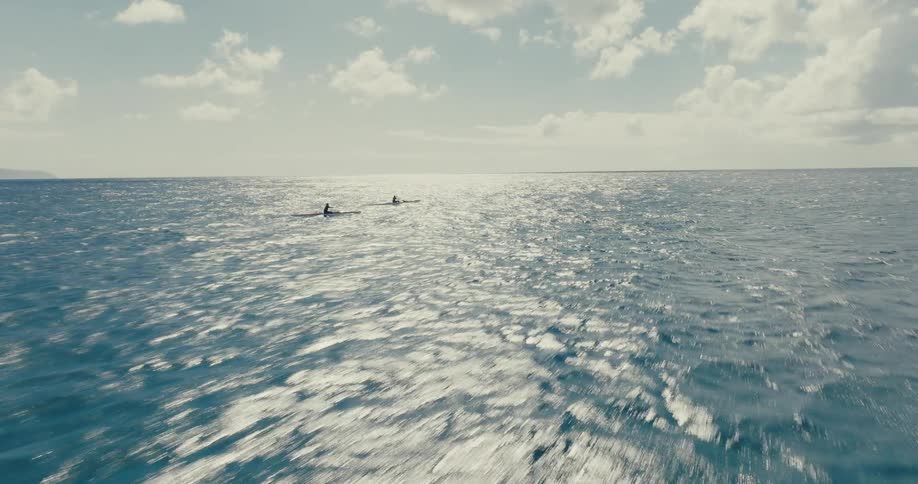 A dramatic aerial shot flies low over the sparkling open ocean, following two people paddling in outrigger canoes