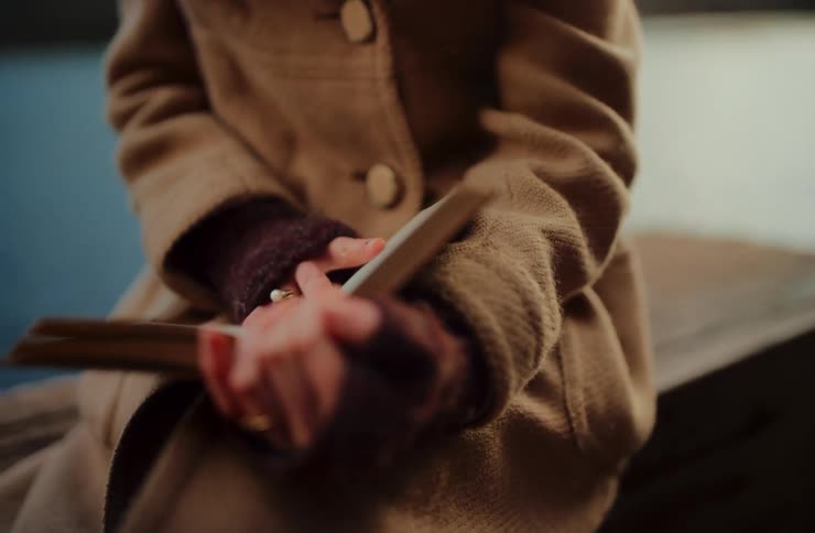 A thoughtful young woman in a warm coat and gloves sits by a tranquil lake and opens a book to read