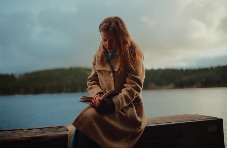 A thoughtful woman in a cozy coat finds a quiet moment to read her book on a wooden pier by a serene lake