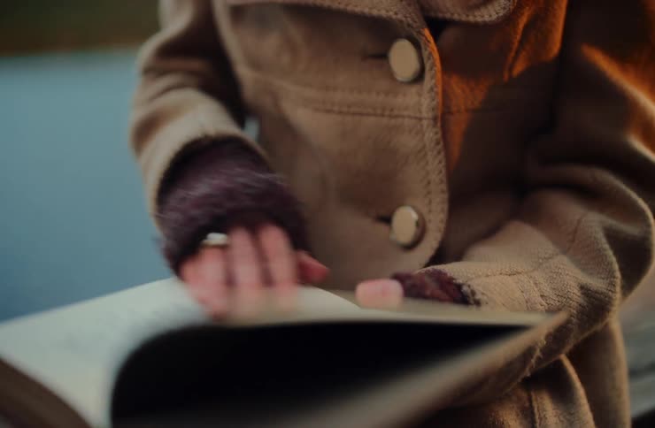 A woman wearing a cozy coat and gloves gently turns the pages of a book in slow motion while sitting outdoors in the warm glow of sunset