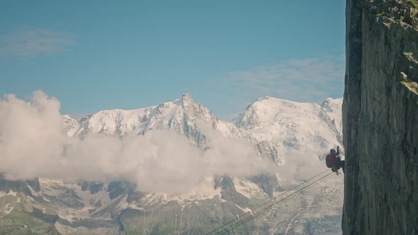 An adventurous climber rappels in slow motion down a sheer rock face with the majestic snow-capped mountains of the Alps in the background