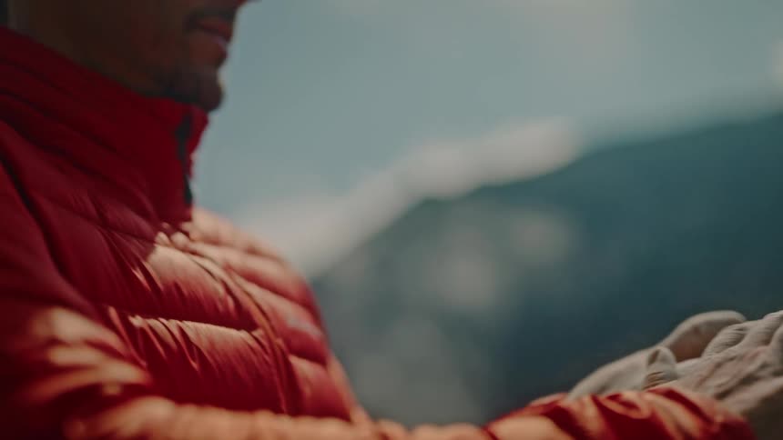 A climber in a red jacket chalks his hands in slow motion, preparing for his ascent as he gazes up at the mountain
