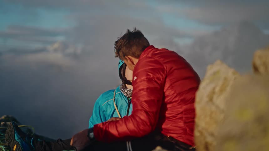 A smiling climber couple share a romantic kiss on a rocky mountain summit above the clouds before preparing their gear