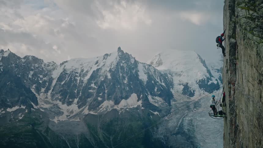 Epic wide shot of two rock climbers ascending a steep cliff face with a breathtaking view of a snow-covered mountain range
