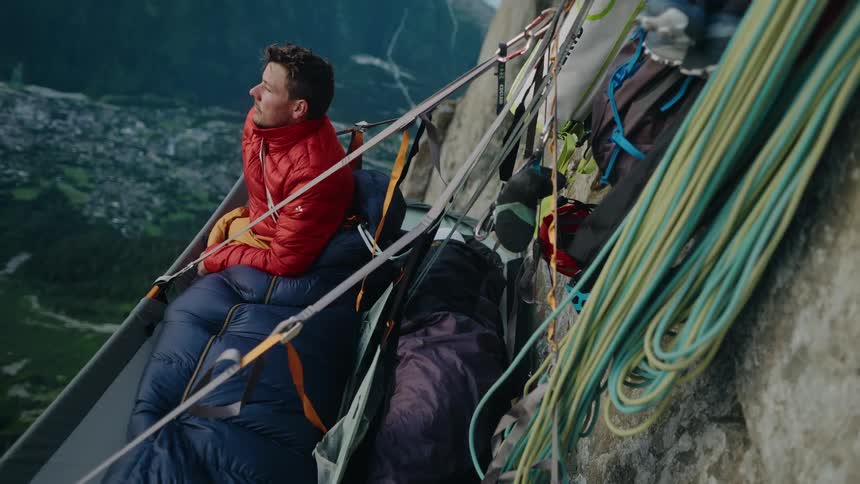 An alpinist in a red jacket wakes up in a portaledge on a sheer mountain face, admiring the vast landscape below