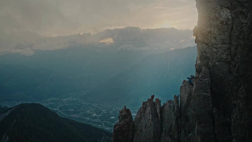 Two rock climbers scale a dramatic mountain peak high above a sprawling valley at sunset