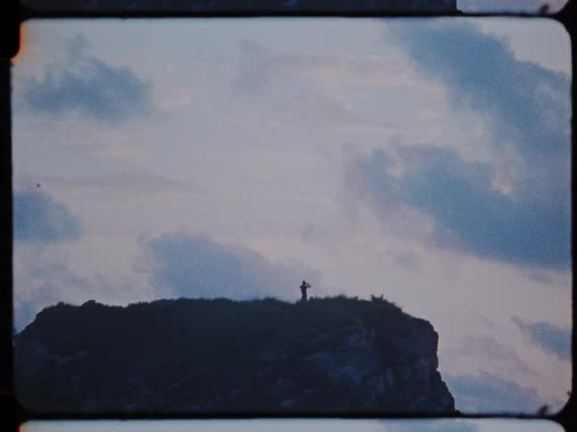 Vintage Super 8 film captures the silhouette of a lone person standing on a clifftop against a vast, cloudy sky