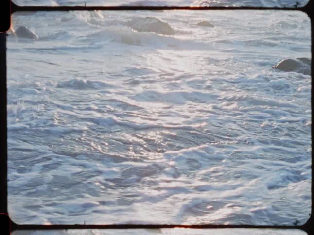 Vintage Super 8 film captures shimmering ocean waves washing over a rocky coastline