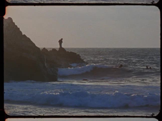 A lone fisherman stands silhouetted on a rocky outcrop as ocean waves crash in this vintage film clip