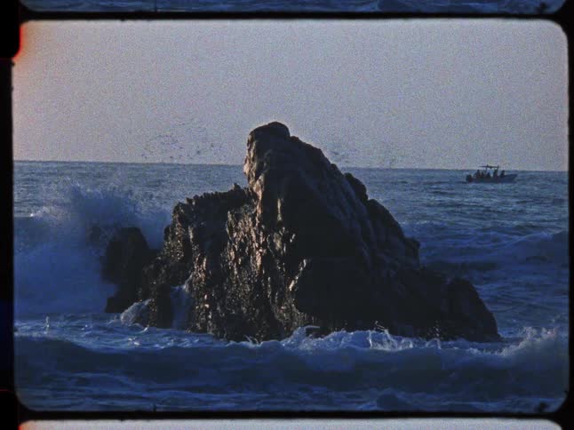 Vintage slow-motion footage shows powerful ocean waves crashing against a large rock as a small boat passes in the distance